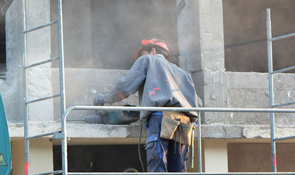 A construction worker stands with his back to the camera, a hammer drill resting on a concrete column, clouded in dust without personal protective equipment.