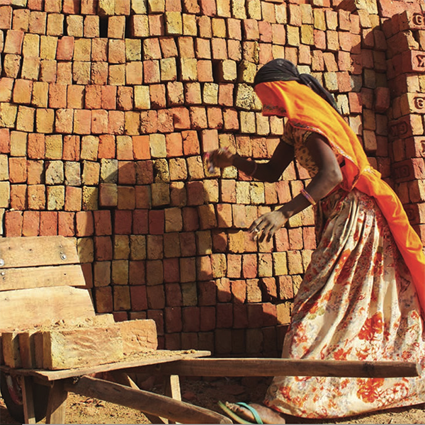 A woman with long, dark hair wearing an orange sari. An orange scarf covers her eyes. She is bending near a neat stack of bricks.