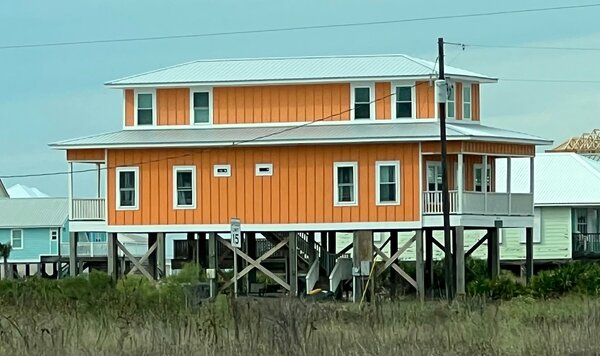 Orange two-story coastal house elevated on timber pilings above beach grass