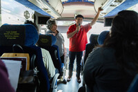  a Latino man in a red shirt and black beret speaks into a microphone on a tour bus while passengers listen.