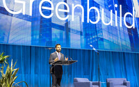 a man with a beard speaks behind a podium with the large word "Greenbuild" as a backdrop.