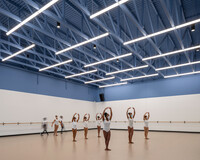 ballerinas of color dance in a studio, wearing white leotards. the floor is a dark earth tone. the ceiling has exposed structural members and is deep blue.
