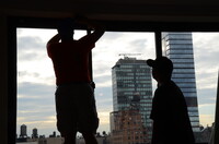 Two men hang shades in an apartment overlooking a city. 