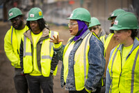 seven people in green hard hats and safety yellow vests. a Black woman talks to someone off camera while the other six people listen.