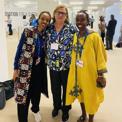 Three women—two black and one white—embrace each other with one arm while smiling at the camera. They are all wearing brightly colored clothes with bold patterns.