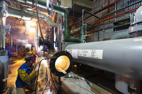 man welding large pipe in mechanical room, sparks flying.