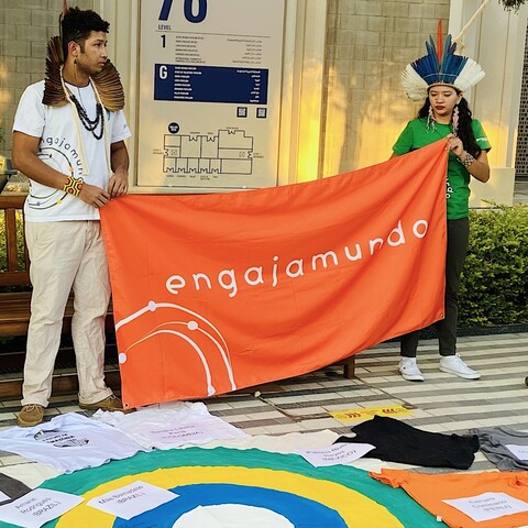 two people, both emblazoned with feathers, hold an orange banner that reads "engajamundo."