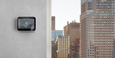 A wall with a small Kaiterra Sensedge monitor with display next to a window overlooking a city skyline.