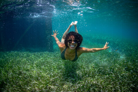 a woman with sunglasses on is under water above sea grass and smiling joyfully at the camera.