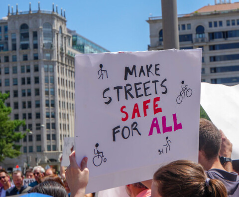 Person at a protest, holding a sign that says, “make streets safe for all,” with drawings of people biking, walking with a cane, walking a dog, and in a wheelchair.