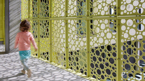A child walking on sun-dappled pavement near a perforated lime green architectural screen.
